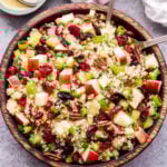 Cranberry Apple Quinoa Salad in wooden bowl with serving spoons. Small bowl of vinaigrette and a dish towel behind the salad.