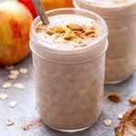 Front facing photo of a cinnamon apple smoothie in glass jar with a second one behind it. Smoothies topped with oats, cinnamon and walnuts. Two apples behind the smoothies.