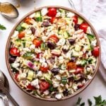Greek Orzo Salad with tomatoes, olives and cucumbers in a round serving bowl with spoons next to it a bowl of vinaigrette and dish towel behind it.