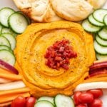 Roasted red pepper hummus on a platter with tomatoes, cucumbers, carrots, and pita bread.