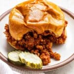 A sloppy joe with a bite taken out of it and pickles on the plate.