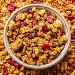 Pumpkin granola in a white bowl with more granola surrounding the bowl.