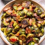 Overhead photo of a white bowl filled with balsamic maple roasted brussels sprouts with bacon. A small bowl of balsamic vinegar with a spoon in it behind the bowl.