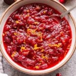 Bourbon Orange Cranberry Sauce in a white serving bowl with a spoon. Half of an orange and a shot of bourbon behind the bowl.