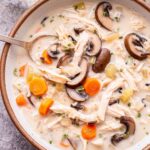 Overhead photo of a white bowl filled with creamy Chicken and Wild Rice Soup with a spoon in the bowl. Soup has sliced mushroom, shredded chicken, wild rice, carrots and celery in it.
