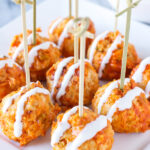 Square white plate filled with buffalo chicken meatballs with toothpicks in them. Meatballs are drizzled with blue cheese dipping sauce.