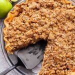 Caramel apple crumb pie in a glass pie plate with a piece cut out and a black spatula in it's place.