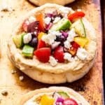 Closeup photo of a Greek pita pizza topped with hummus, tomatoes, feta, red onion, cucumber and olives.