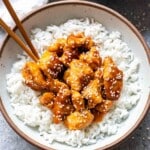 Sesame chicken on top of rice in a white bowl with chopsticks.