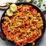 Chicken Fajitas in a skillet with two lime halves. Tortillas are behind the skillet.
