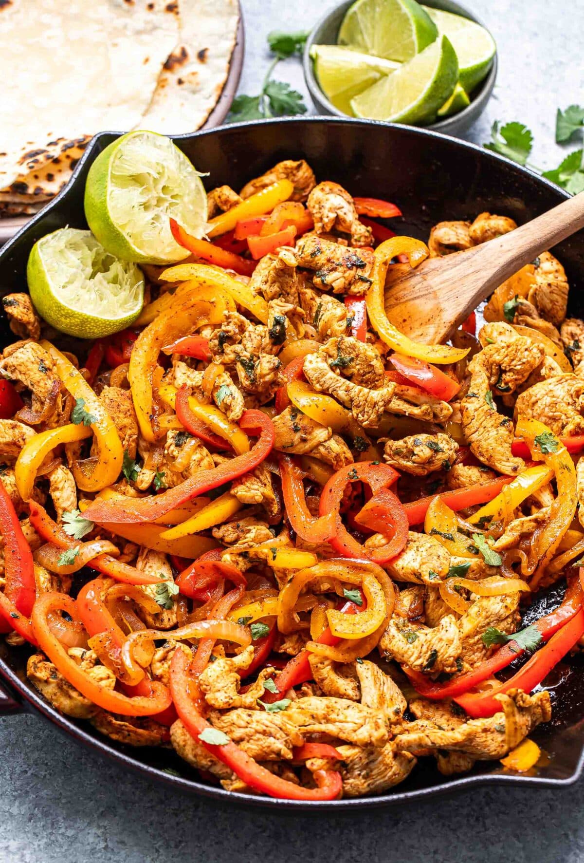 Chicken Fajitas in a skillet with a wooden spoon and two lime halves.