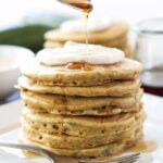 A stack of Zucchini Bread Pancakes topped with Maple Cream Cheese and syrup being spooned over them.