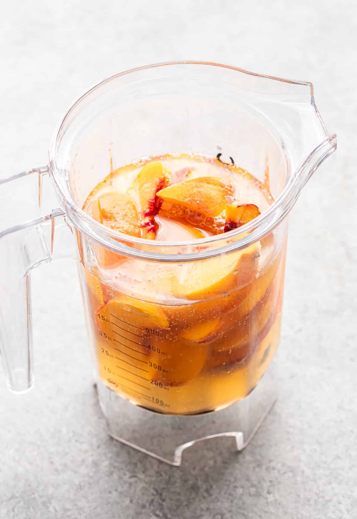 A clear pitcher filled with sliced peaches and liquid, reminiscent of a refreshing Bourbon Peach Slush, sits on a light gray surface.