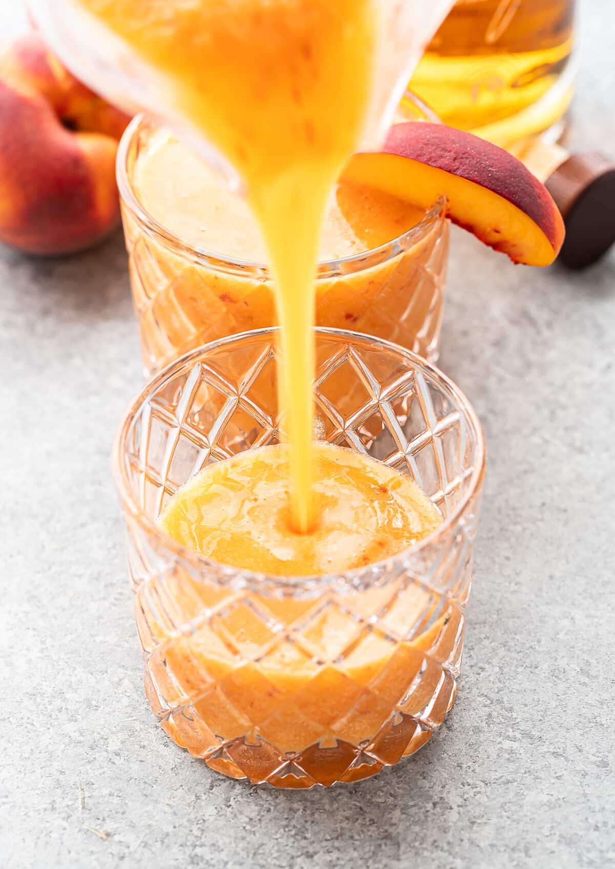 A thick, peach-colored Bourbon Peach Slush is being poured into a textured glass, with another filled glass and fresh peaches in the background.