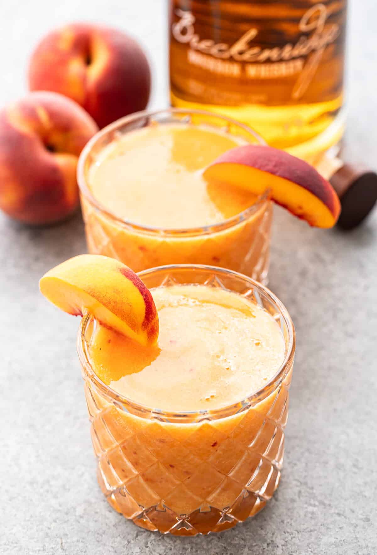 Two glasses of peach-colored Bourbon Peach Slush cocktails garnished with peach slices sit on a gray surface. A bottle of whiskey and two whole peaches are in the background.
