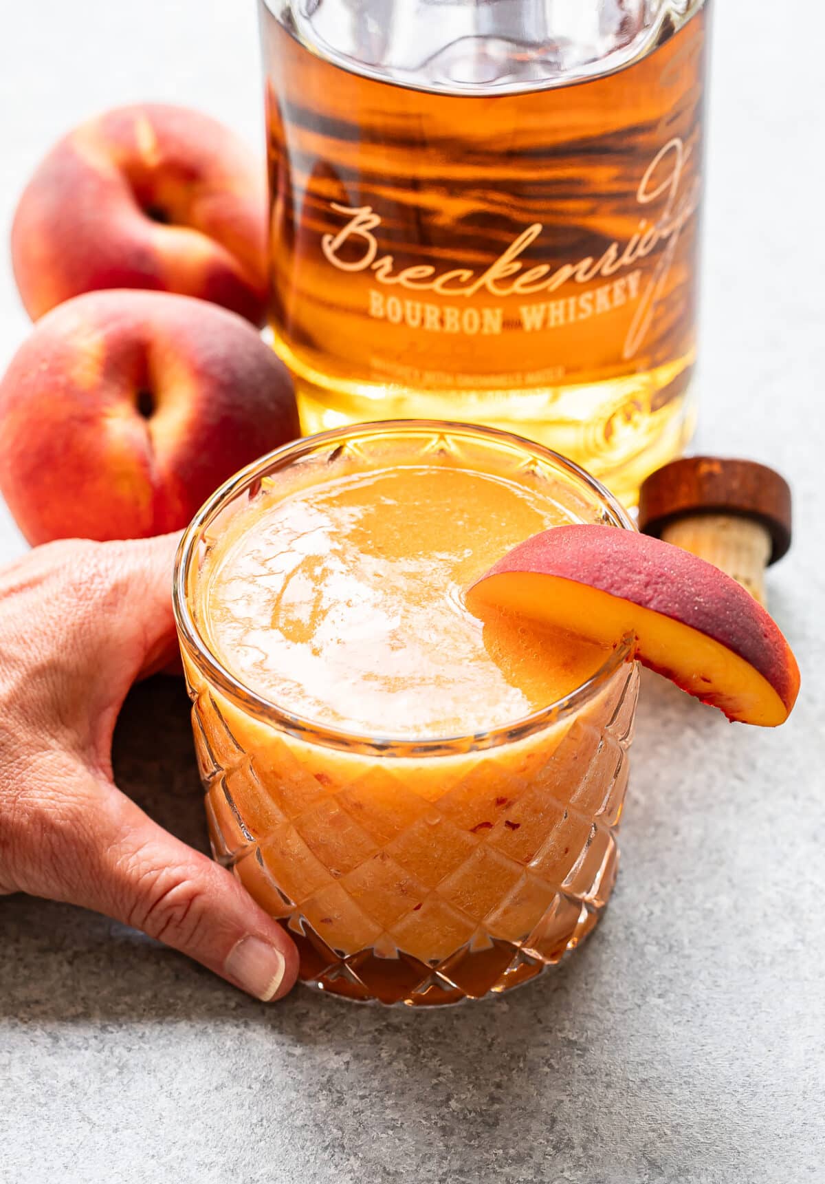 A hand holds a glass of Bourbon Peach Slush garnished with a peach slice, next to fresh peaches and a bottle of Breckenridge bourbon whiskey on a gray surface.