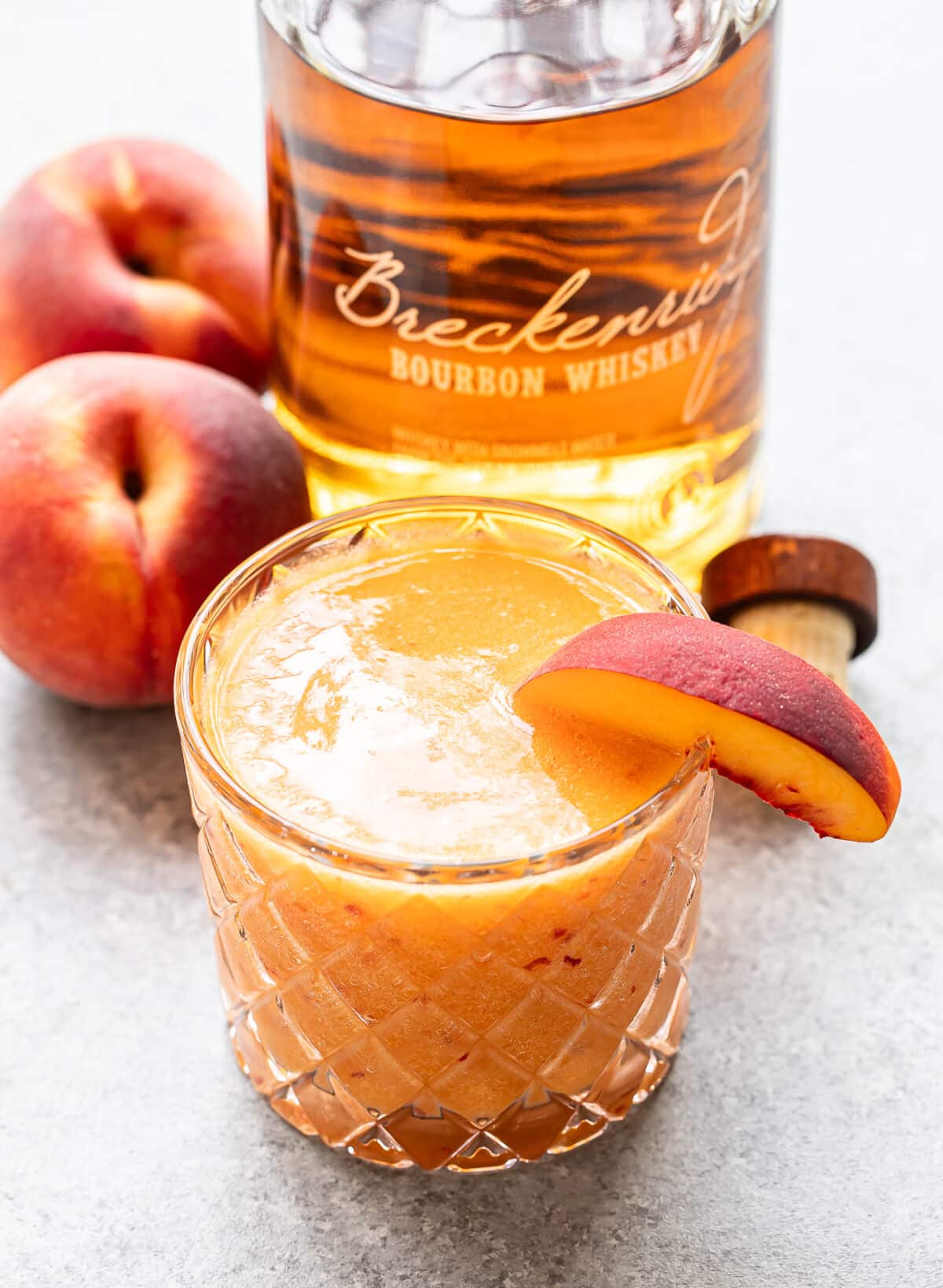 A glass of Bourbon Peach Slush, garnished with a peach slice, sits next to two whole peaches and a bottle of bourbon whiskey on a gray surface.