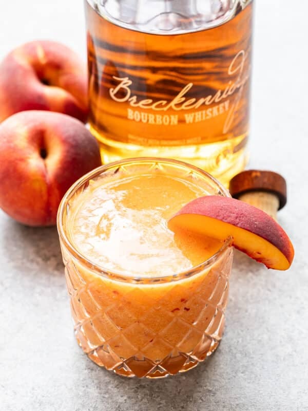 A glass of Bourbon Peach Slush, garnished with a peach slice, sits next to two whole peaches and a bottle of bourbon whiskey on a gray surface.
