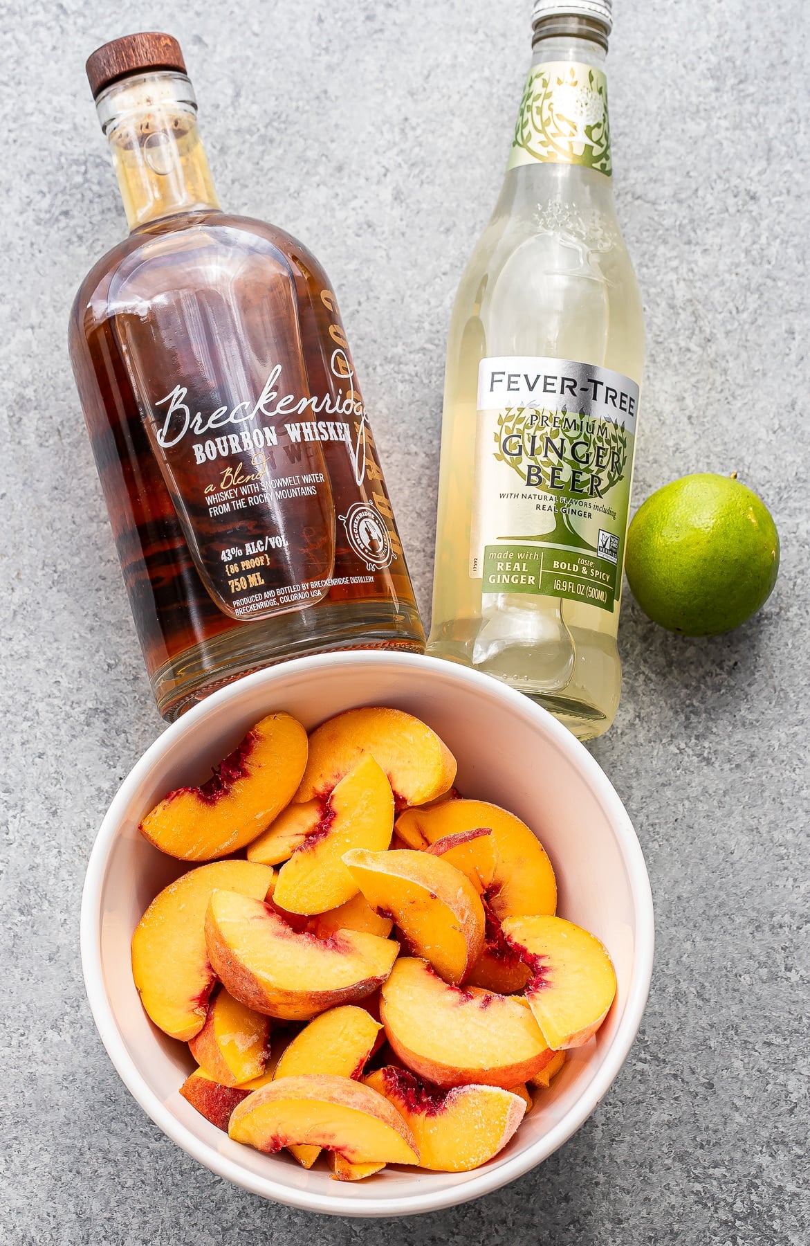 A bowl of sliced peaches sits next to a bottle of bourbon, a bottle of ginger beer, and a whole lime on a gray countertop—perfect ingredients for crafting a refreshing Bourbon Peach Slush.