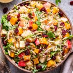 Overhead photo of Harvest quinoa salad with butternut squash, apples, dried cranberries, pepitas, spinach, and toasted almonds in a white serving bowl. A jar of cider vinaigrette behind the bowl.