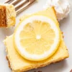 Lemon Greek yogurt cheesecake bar on a plate topped with a lemon slice. A corner of it is cut off and on a fork.