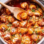 Enchilada Meatballs topped with melted cheese in a stainless steel pan with a wooden serving spoon in the pan.