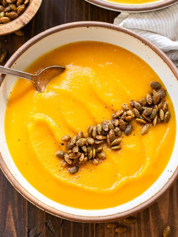 A bowl of roasted butternut squash and pumpkin soup topped with roasted pumpkin seeds and a metal spoon.