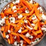 Honey Roasted Carrots with goat cheese and thyme leaves in a white serving bowl with two serving spoons next to the bowl.