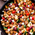 Greek Chicken and Vegetables in a black cast iron skillet with a bowl of feta behind it. Skillet includes chicken, zucchini, tomatoes, kalamata olives and feta crumbles.