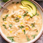 Overhead photo of Green Chile Chicken Enchilada Soup in a white bowl with a spoon. Shredded chicken, white beans, corn, green chiles and cilantro are in the soup. Melted cheese and avocado slices on top. A small bowl of shredded cheese is behind the bowl of soup.