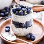 Blueberry Lemon Overnight Oats in a glass jar on top of a wooden plate with a spoon on the plate. Oats are topped with fresh blueberries and lemon zest.