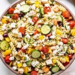 Greek Chicken Pasta Salad in a white serving bowl with serving spoons and a bowl of vinaigrette behind it.