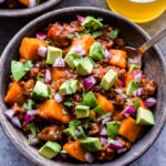 Slow Cooker Bison Sweet Potato Chili in grey bowl with spoon. Topped with avocado, red onions and cilantro.