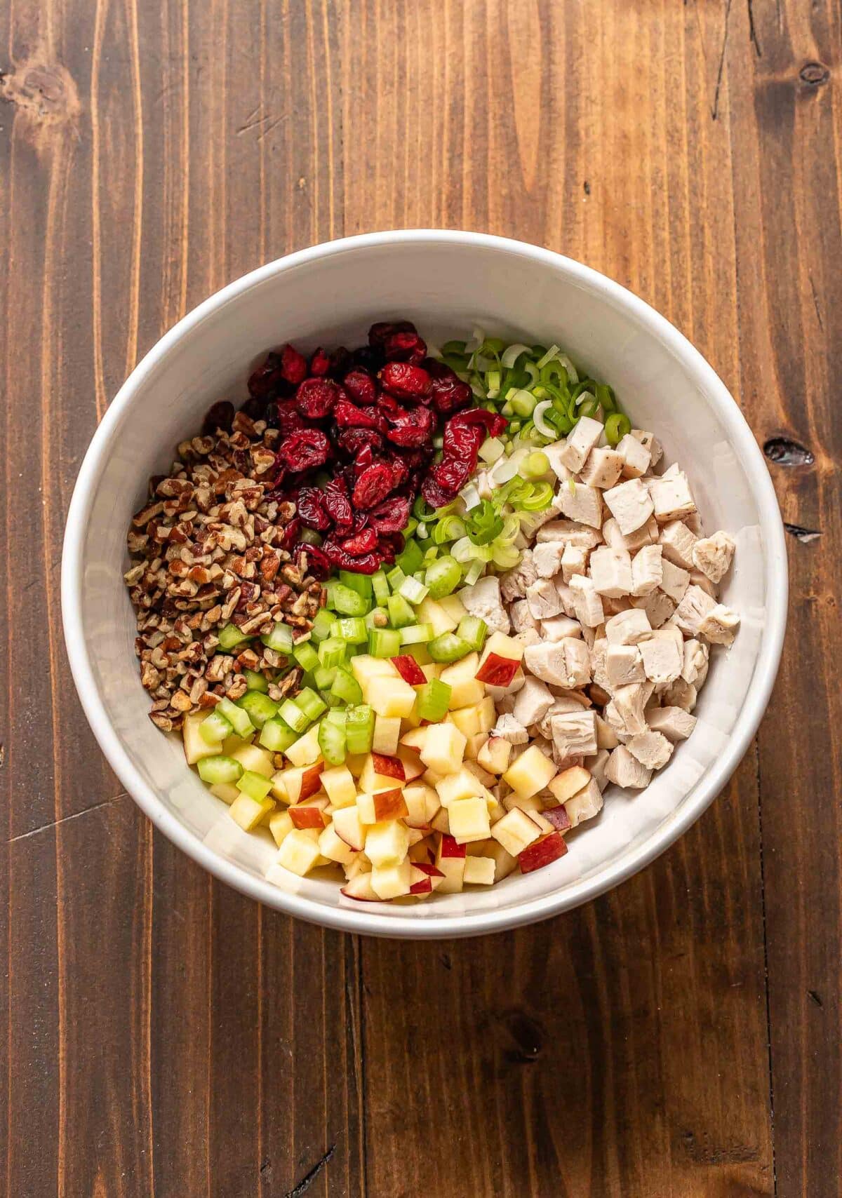 chicken, celery, dried cranberries, apple, celery, and pecans in a white mixing bowl.