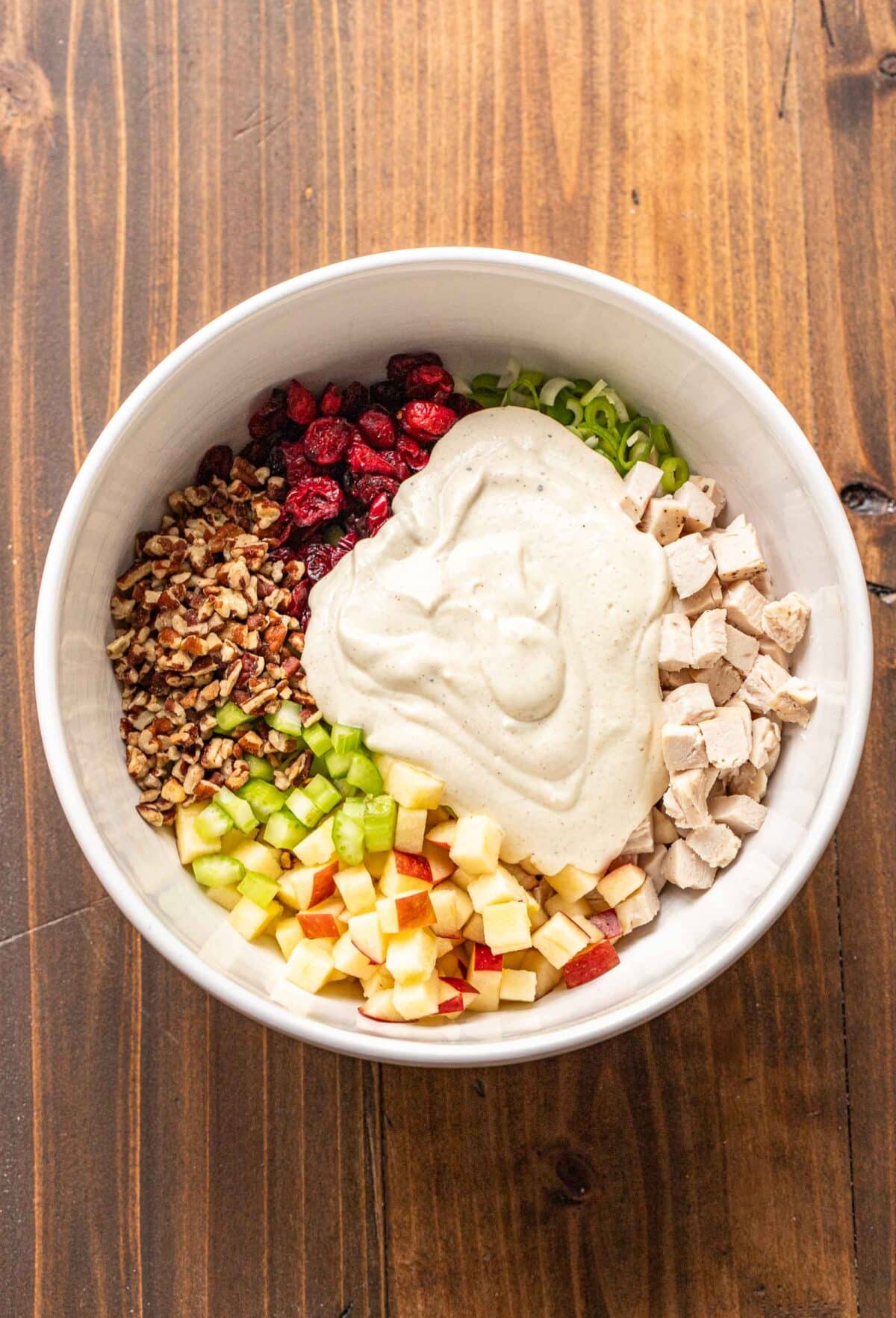 chicken, celery, dried cranberries, apple, celery, pecans, with a creamy dressing on top in a white mixing bowl.