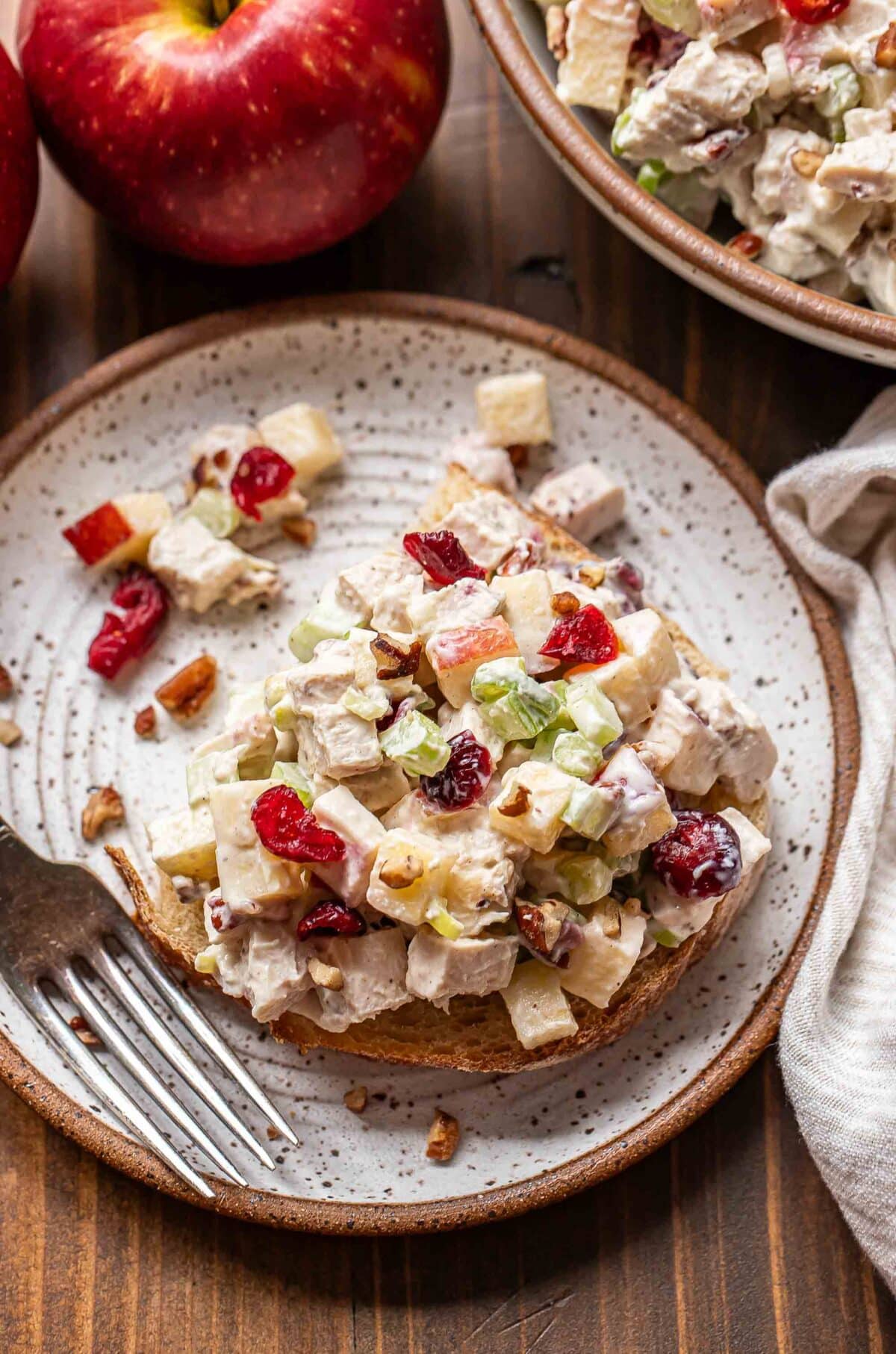 Chicken salad on top of a piece of bread on a white plate with a fork. An apple and bowl of chicken salad is behind the plate.