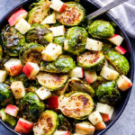 Brussels sprouts and apple salad in black serving bowl with spoons. Maple mustard dressing behind the bowl.