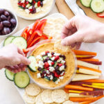 A platter with crackers, sliced carrots, red bell pepper strips, cucumber, and a bowl of hummus topped with feta, olives, and red peppers. Two hands are dipping a cracker and a cucumber slice into the hummus.