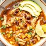 Chicken Tortilla Soup in a white soup bowl with a spoon in it and avocado slices and lime wedges on top.