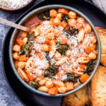 Chickpea pasta fagioli in a bowl with a spoon. The bowl is on a black plate with slices of baguette. A bowl of parmesan cheese is in the background.
