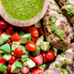 Grilled Chicken with Tomato Avocado Salad drizzled with green chimichurri sauce. Overhead, closeup photo.