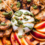 Overhead photo of round platter with grilled peach slices, baguette slices and burrata in the center, cut open. Drizzled with pesto and balsamic glaze.