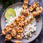 Overhead photo of Coconut Lime Chicken Skewers over rice with lime wedges on gray plate.