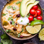 Overhead photo of Southwest Chicken and Rice Soup in bowl with a spoon topped with avocado, tomatoes, lime wedges, sour cream and cilantro.