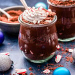 Peppermint Chocolate Pudding Parfaits topped with coconut whipped cream and crushed candy canes in jars with spoons in them.