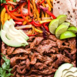 Steak Fajitas on a rimmed sheet pan with tortillas, avocado slices, lime wedges and cilantro