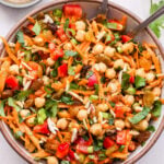 Overhead photo Moroccan Chickpea Salad with carrots, red bell pepper, golden raisins, almonds, cilantro and mint in white serving bowl. Small bowl of vinaigrette behind the salad.
