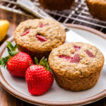 Two Strawberry Banana Muffins on a round white plate with a butter knife and two strawberries.