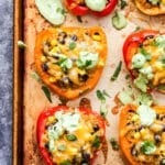 Six Mexican Quinoa Stuffed Peppers on a rimmed baking sheet with a spoon covered in avocado sauce.