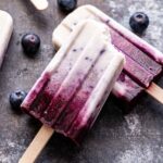 Two blueberry coconut milk popsicles. One propped up on the other with a bite taken out of it. Fresh blueberries scattered around them.